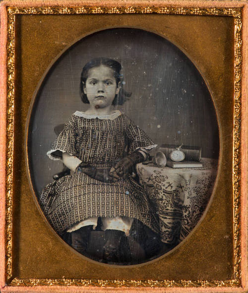 School girl seated at a table with two books, a bell, and a pocket watch
