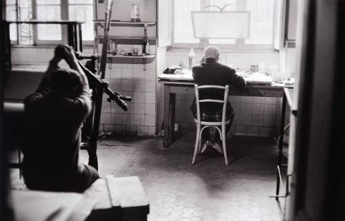 Jacqueline sits on an empty box in basement of villa, which Picasso converted into engraver's printing plant, while he etches the copper plates of the bullfight series. Picasso works by the window revetment.