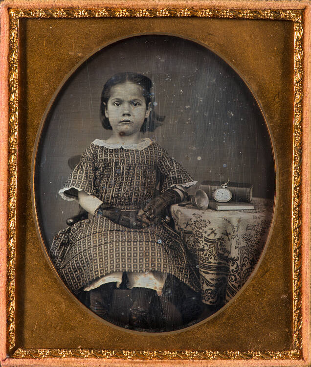 School girl seated at a table with two books, a bell, and a pocket watch