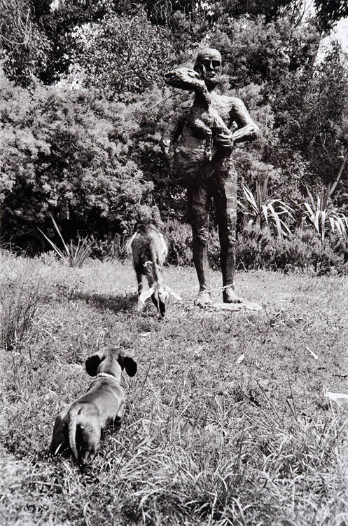 Lump, the dachshund, meets the pet goat for the first time, in garden. Goat is tethered to bronze statue, "Man with a Sheep" or "Shepherd Carrying a Lamb" (1944). Over seven feet tall.